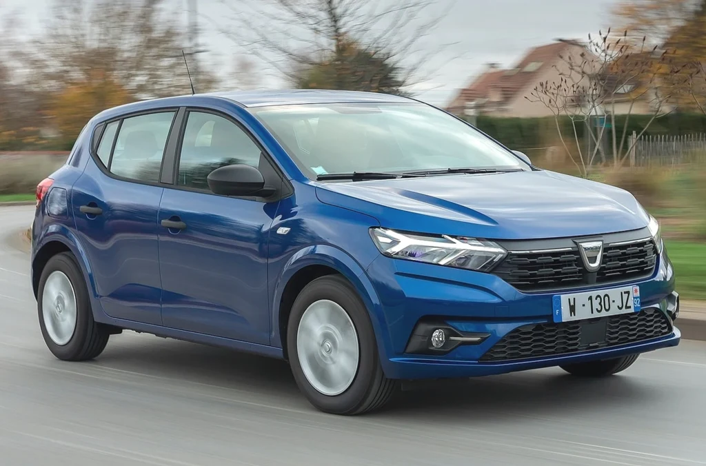 Model, wyceniony na 61,3 tys. zł, od lat należy do liderów sprzedaży w Unii Europejskiej. Niebieska Dacia Sandero w ruchu na asfaltowej drodze, w tle jesienne drzewa i zabudowania mieszkalne po prawej stronie.