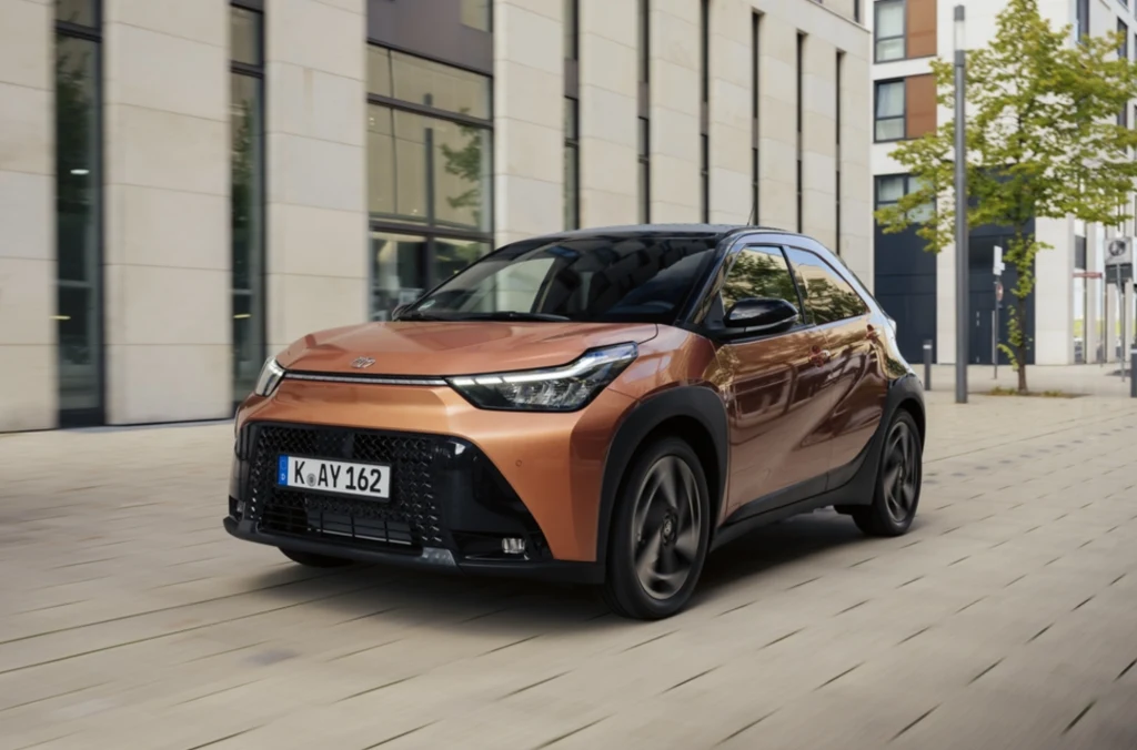 Tuż za podium znajduje się Toyota Aygo X. Nowoczesny, brązowy samochód osobowy typu crossover poruszający się miejską ulicą na tle wysokich, nowoczesnych budynków z dużymi oknami.