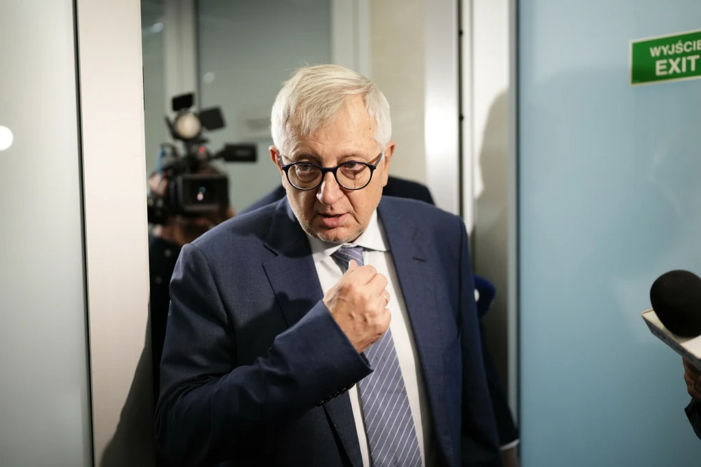 Marek Siwiec Starszy mężczyzna w okularach i garniturze znajduje się we wnętrzu budynku, wykonuje gest zaciśniętej pięści, w tle widoczna kamera oraz znak EXIT.