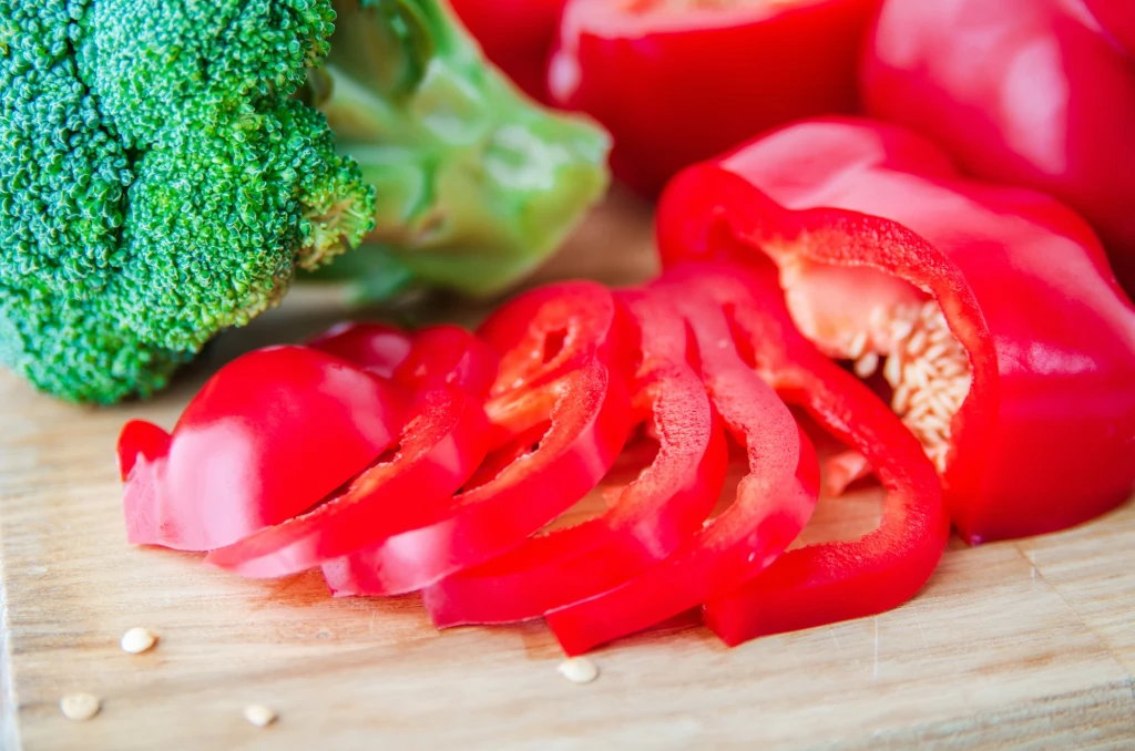 Papryka i brokuły to warzywa będące dobrym źródłem witaminy C Pokrojona czerwona papryka oraz świeży brokuł leżące na drewnianej desce, w tle widoczne są kawałki czerwonej papryki.