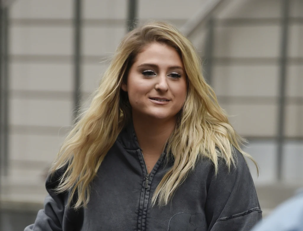 Młoda kobieta z długimi blond włosami ubrana w ciemną bluzę, stoi na zewnątrz na tle rozmytego betonowego budynku i metalowej barierki.