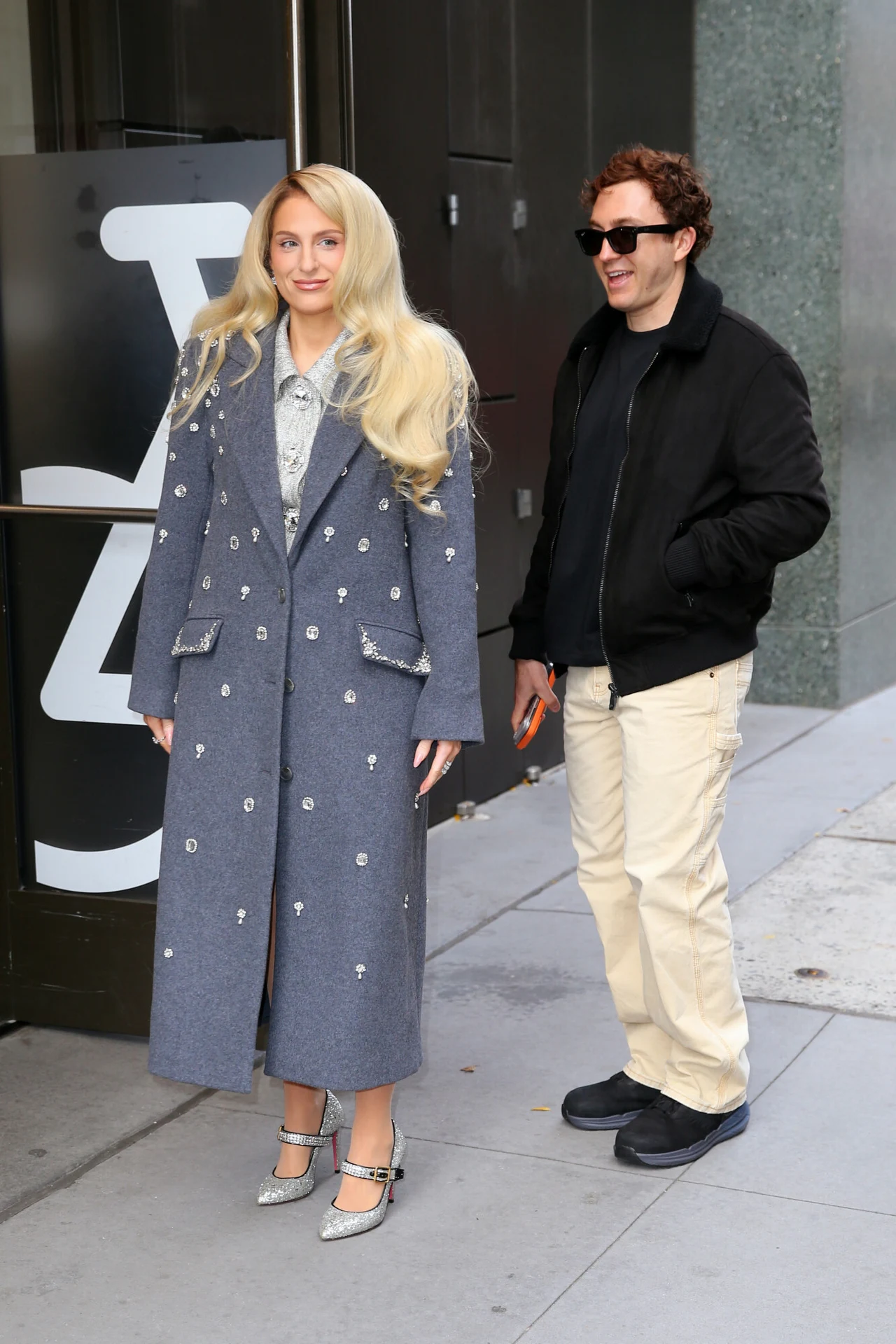 Kobieta w długim szarym płaszczu z ozdobami stoi na chodniku obok uśmiechniętego mężczyzny w czarnej kurtce, jasnych spodniach i okularach przeciwsłonecznych. Oboje znajdują się w przestrzeni miejskiej.