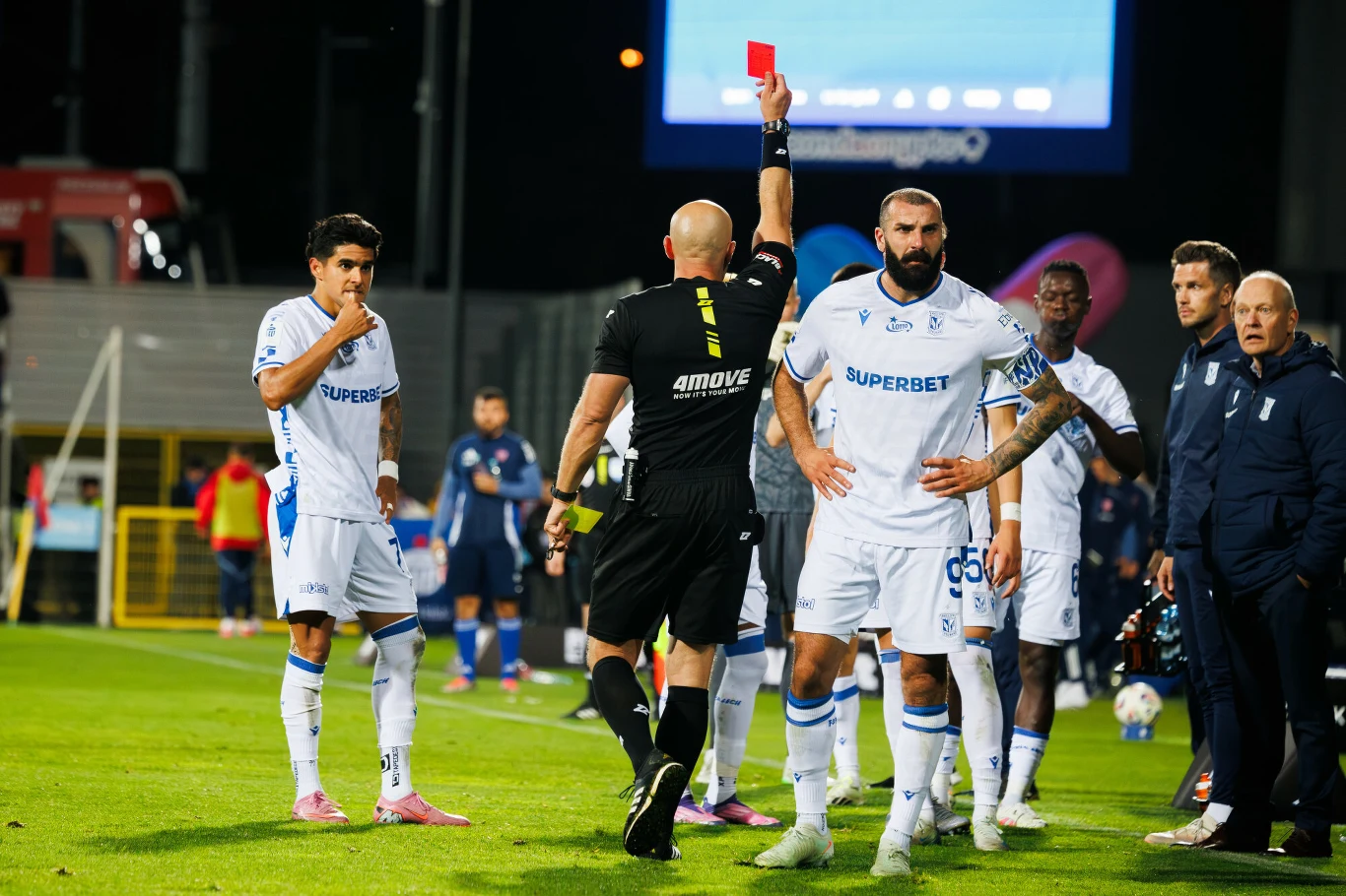 Szymon Marciniak i pozostali sędziowie Ekstraklasy bedą tłumaczyli swoje decyzje Sędzia pokazuje czerwoną kartkę jednemu z piłkarzy ubranych w białe stroje podczas meczu piłkarskiego, wokół niego stoją inni zawodnicy i członkowie sztabu, na tle widoczne trybuny i elementy stadionu.
