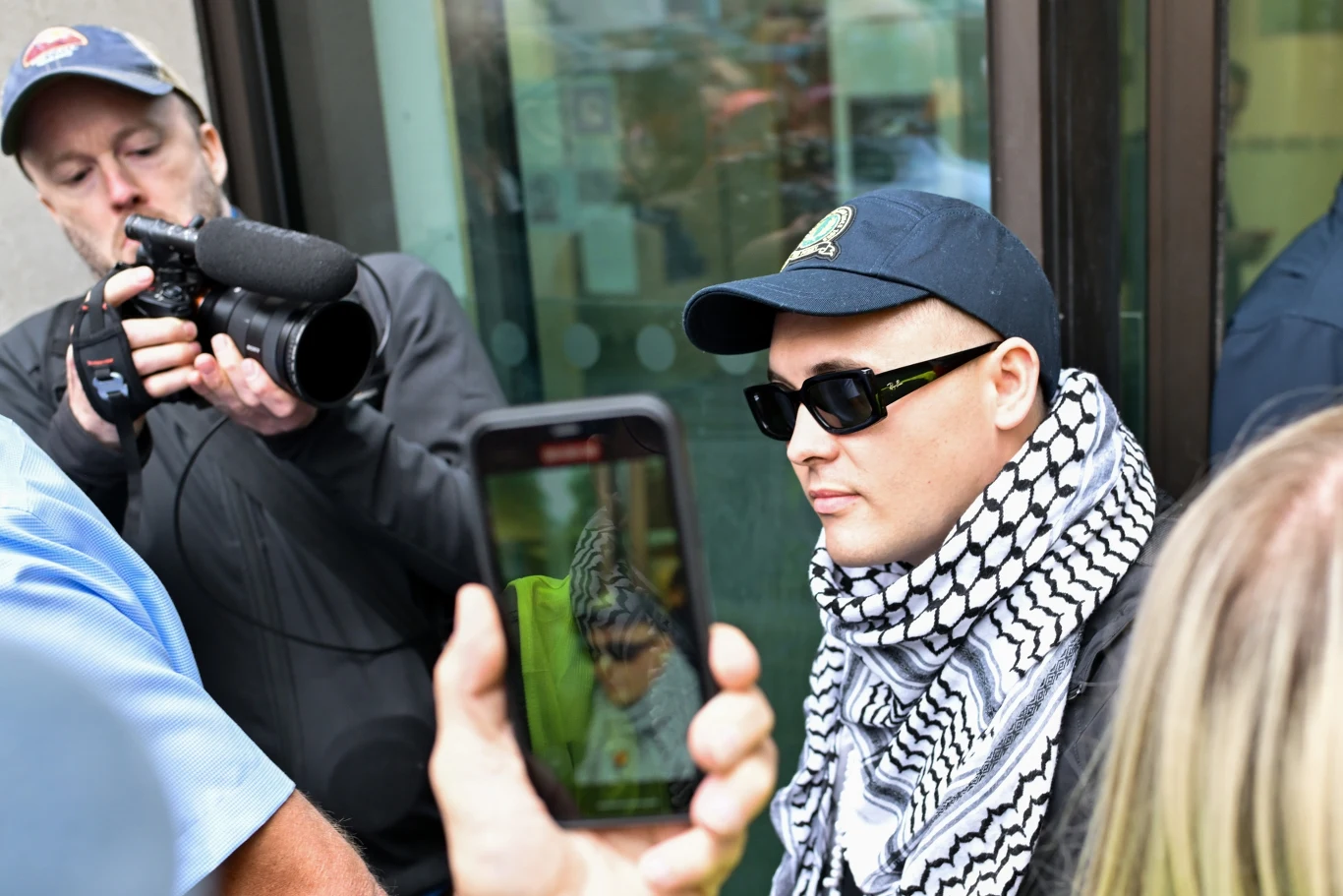 Liam Óg Ó Hannaidh (Mo Chara) z grupy Kneecap Osoba w czapce z daszkiem i okularach przeciwsłonecznych, owinięta szalikiem w czarno-białą kratę, stoi w otoczeniu innych osób; jedna z nich robi zdjęcie smartfonem, a druga rejestruje zdarzenie profesjonalnym aparatem.