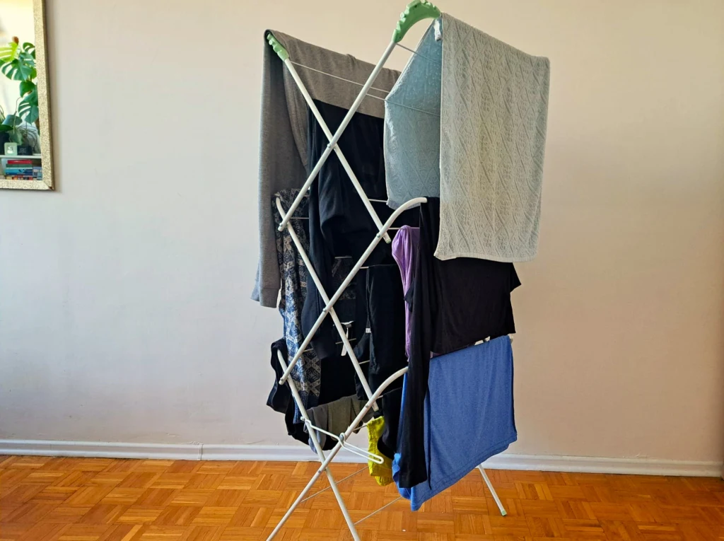 Rozstawiona suszarka na ubrania z kilkoma elementami garderoby i ręcznikiem rozwieszonymi do wyschnięcia, ustawiona na parkiecie przy jasnej ścianie.