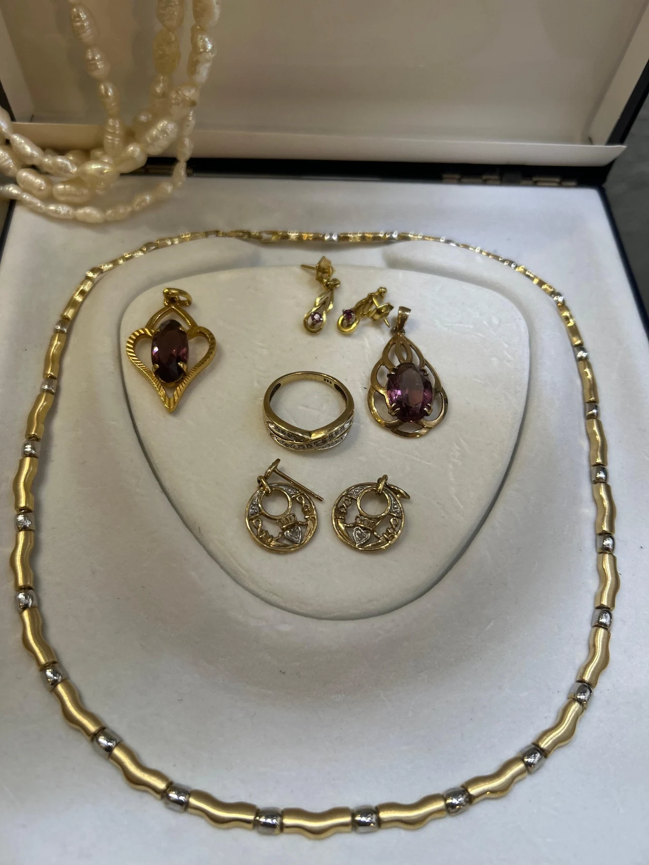 Zestaw biżuterii składający się z naszyjnika, pierścionka, dwóch par kolczyków oraz broszek, umieszczonych w eleganckim pudełku. Dominują złote i srebrne elementy, część ozdób ma kamienie w odcieniach fioletu. W tle widoczny jest sznur pereł.
