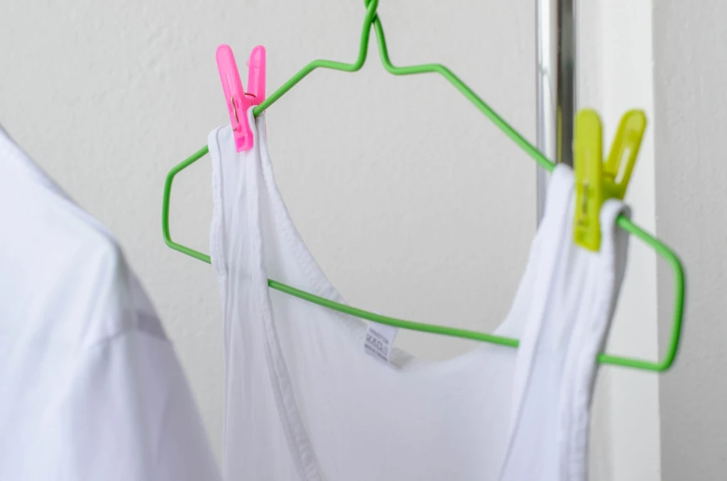 Biały podkoszulek zawieszony na zielonym wieszaku, przymocowanym do metalowego drążka za pomocą różowej oraz żółtej klamerki do bielizny.