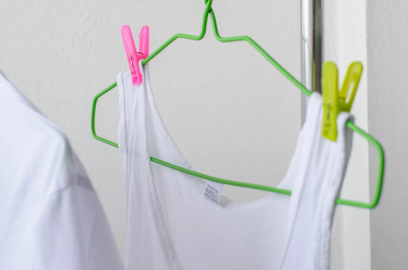 Biały podkoszulek zawieszony na zielonym wieszaku, przymocowanym do metalowego drążka za pomocą różowej oraz żółtej klamerki do bielizny.