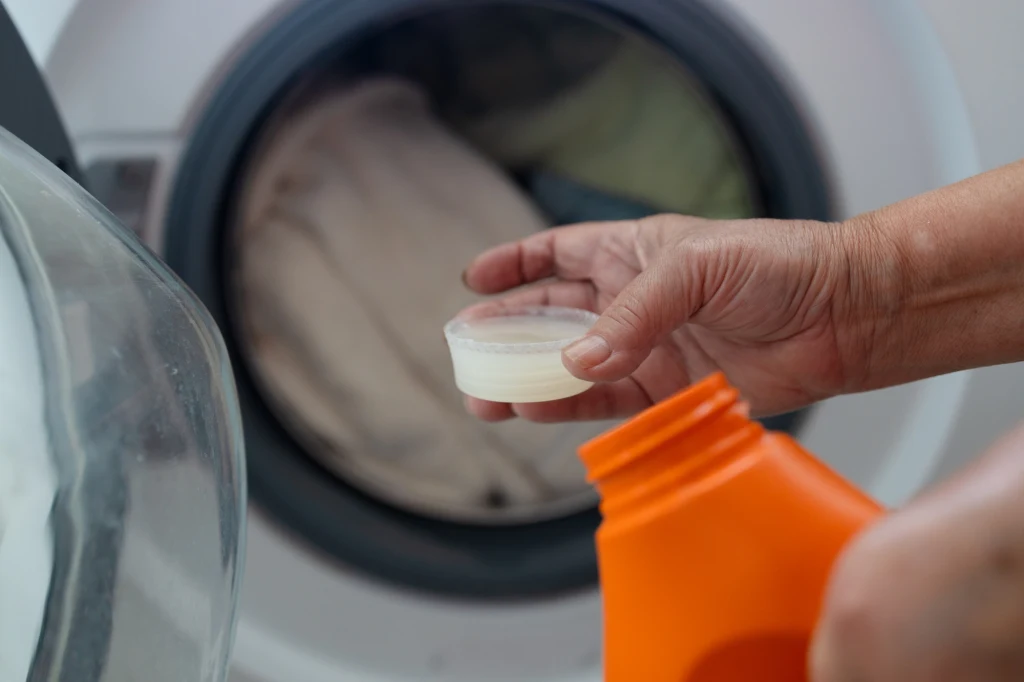 Ręka trzymająca miarkę z płynnym detergentem na tle uchylonych drzwi pralki z praniem w bębnie.