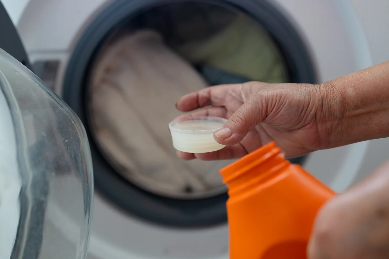 Ręka trzymająca miarkę z płynnym detergentem na tle uchylonych drzwi pralki z praniem w bębnie.