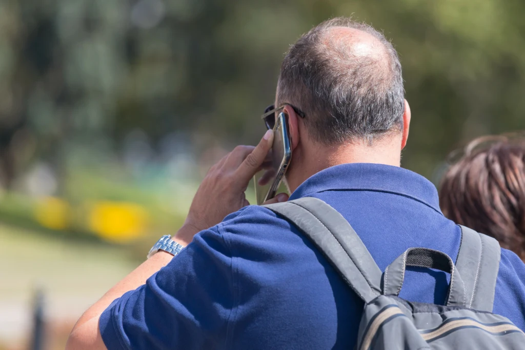 Mężczyzna z plecakiem ubraną w niebieską koszulkę rozmawia przez telefon komórkowy na świeżym powietrzu, w tle rozmyta zieleń i druga osoba częściowo widoczna.
