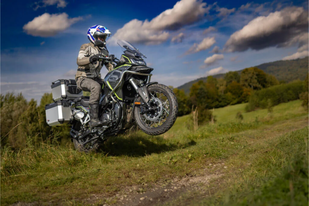 Motocyklista w pełnym stroju ochronnym wykonuje skok terenowy na motocyklu adventure, w tle zielone łąki i drzewa oraz widoczne pagórki pod niebem z chmurami.