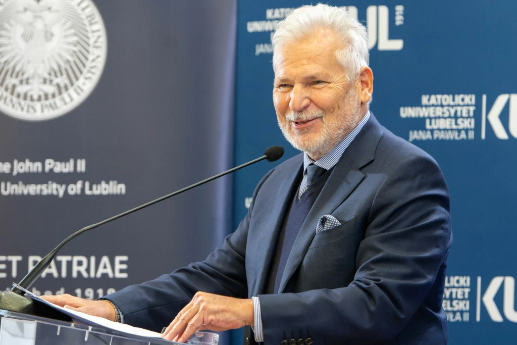 Starszy mężczyzna w eleganckim garniturze uśmiecha się i przemawia przy mównicy, w tle widoczne logo Katolickiego Uniwersytetu Lubelskiego Jana Pawła II.