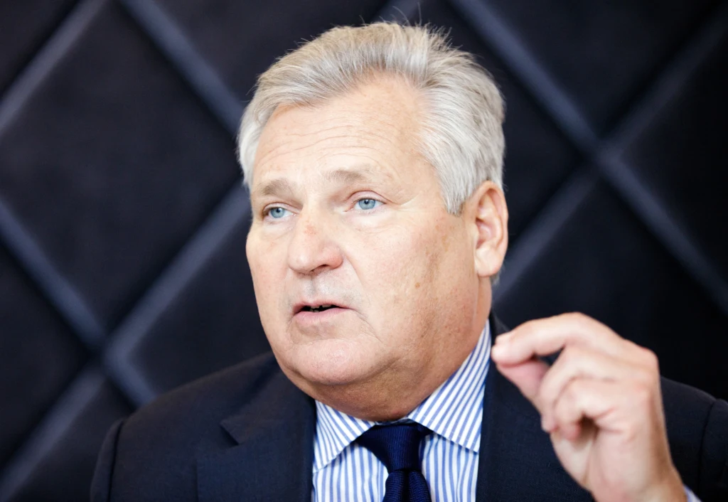 Starszy mężczyzna w eleganckim garniturze i niebieskim krawacie wykonuje gest ręką na tle ciemnej ściany z geometrycznym wzorem. Wyraz twarzy sugeruje zaangażowanie w rozmowę lub wypowiedź.