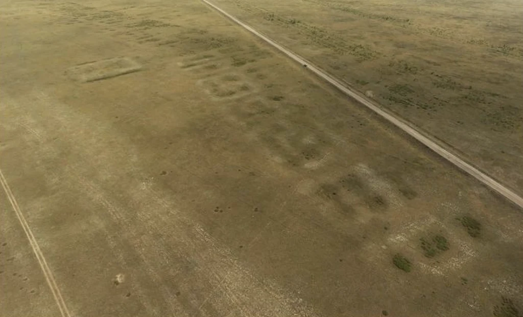 Rozległa równina z widocznymi geometrycznymi kształtami i wzorami w trawie, sugerującymi dawną obecność struktur lub działalności człowieka, oraz biegnąca przez teren prosta droga.