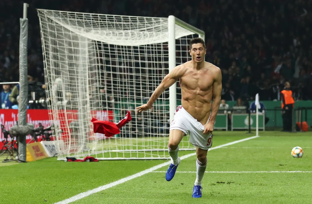 Robert Lewandowski celebrujący gola w meczu Bayernu Monachium w RB Lipsk. Sportowe zdjęcie roku w 2019 roku w Niemczech Rozemocjonowany piłkarz biegnący boisku bez koszulki, w tle bramka piłkarska oraz tłum na trybunach, tuż obok porzucona czerwona koszulka.