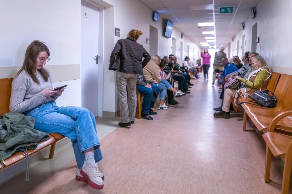 Jedno krótkie L4 w roku nie ma praktycznie znaczenia dla emerytury. Długi korytarz w przychodni lub szpitalu, na ławkach po obu stronach siedzi wielu ludzi czekających w kolejce do lekarza; jedna młoda kobieta siedzi osobno z tabletem, reszta osób zajmuje się sobą, część rozmawia, inni patrzą przed siebie.