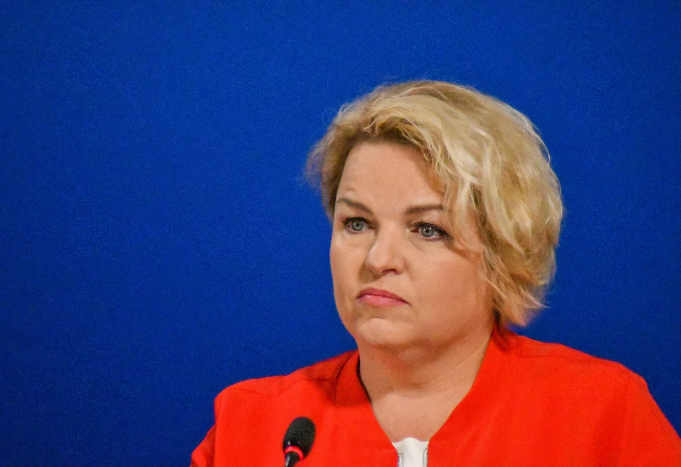 Kobieta o blond włosach w czerwonym ubraniu siedzi przed niebieskim tłem, patrząc poważnie w stronę kamery, przed nią znajduje się mikrofon.