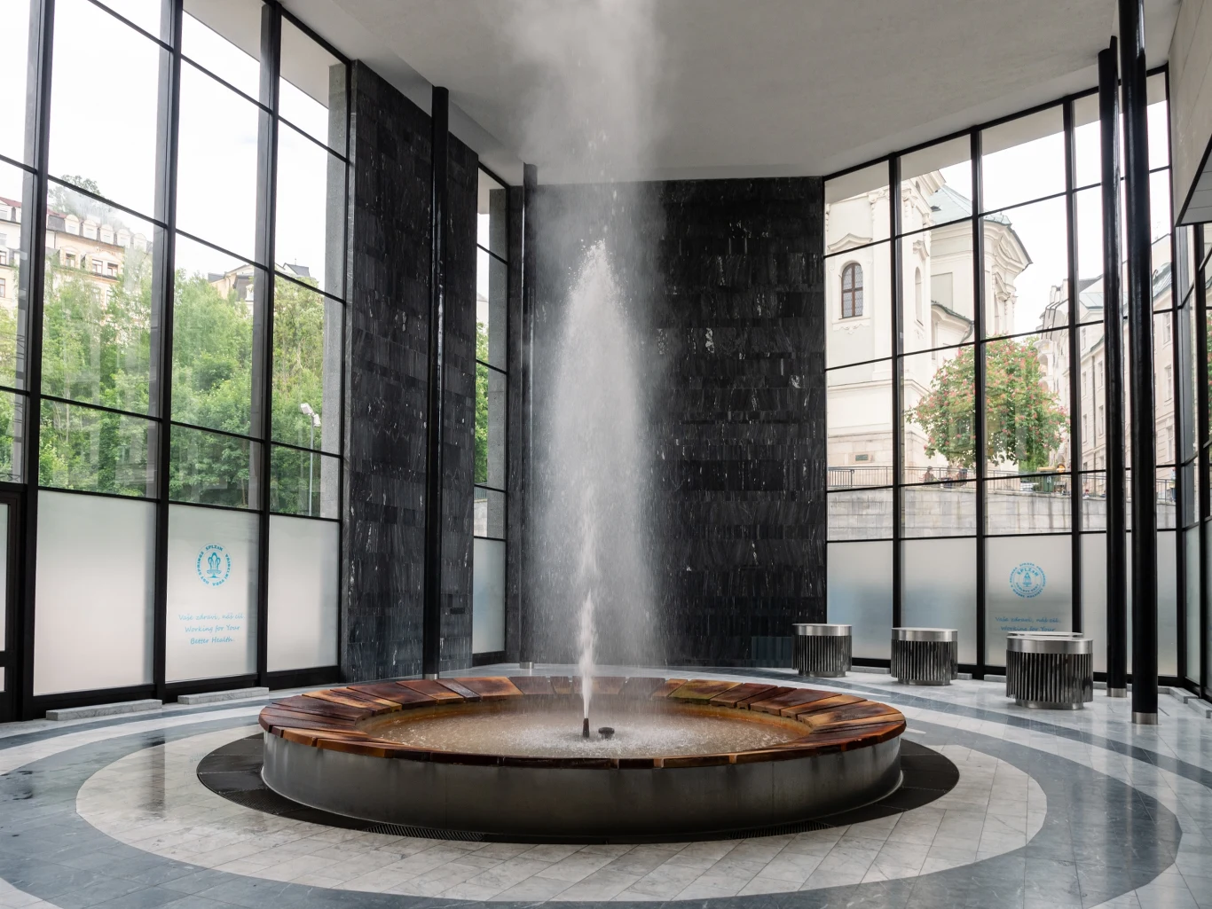 Wytryskujący na wysokość 12 m gejzer Vřídlo to symbol czeskiego uzdrowiska Fontanna mineralna w nowoczesnym, przeszklonym wnętrzu sanatorium, z okrągłym basenem i silnym strumieniem wody skierowanym ku górze, otoczona jasnymi ścianami i dużymi oknami z widokiem na zieleń i klasyczne budynki miasta.