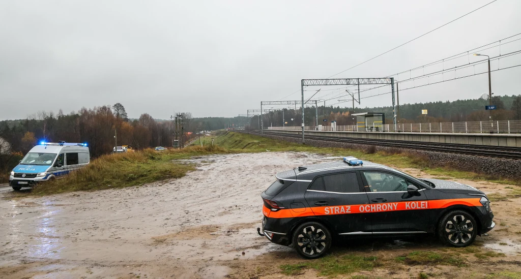 Samochód Straży Ochrony Kolei zatrzymany na błotnistym terenie w pobliżu nasypu kolejowego, w oddali widoczny radiowóz policyjny z włączonymi światłami, tory kolejowe biegnące przez las pod chmurami na ponurym niebie.