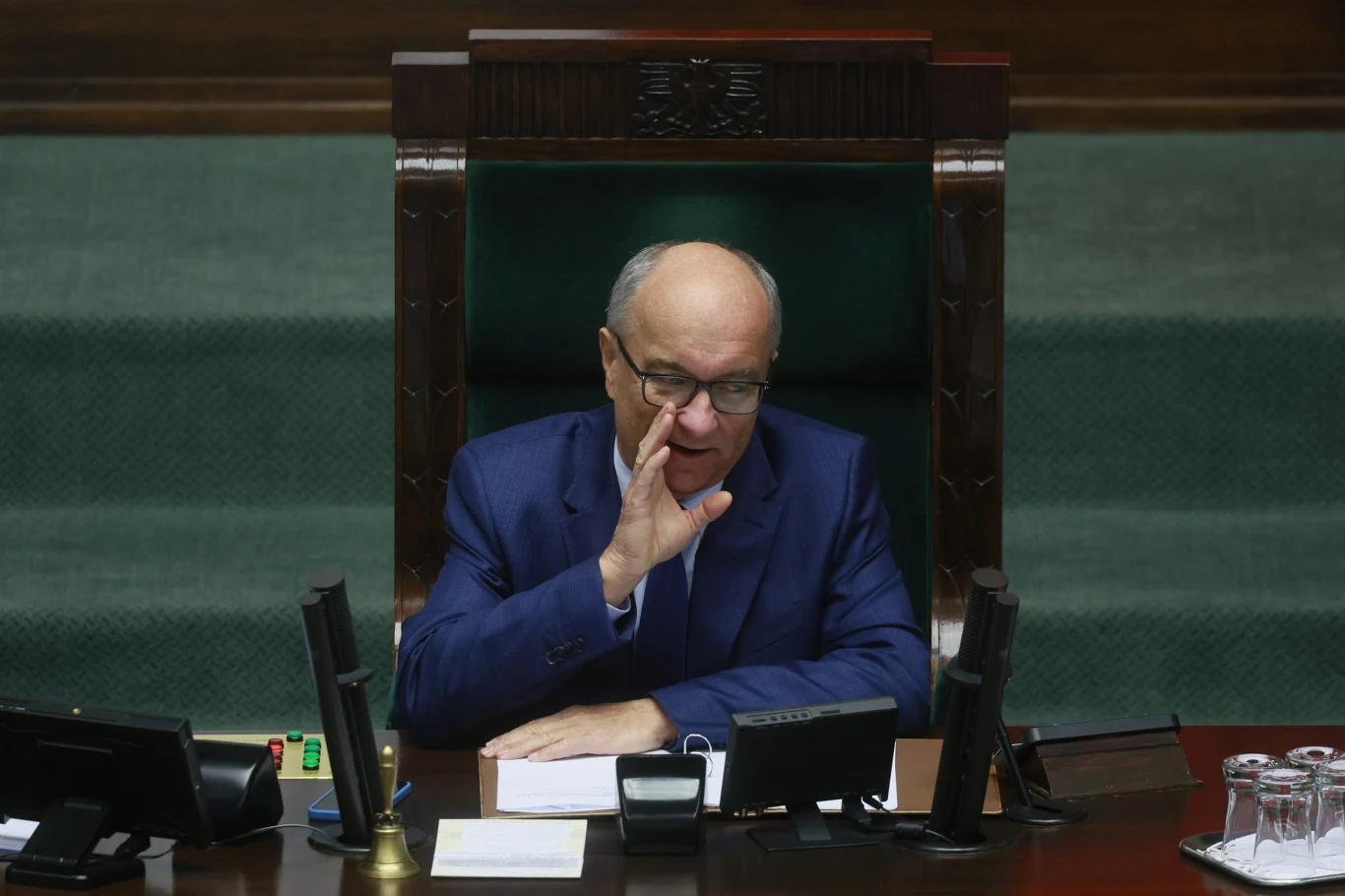 Włodzimierz Czarzasty nowym marszałkiem Sejmu Mężczyzna w garniturze siedzi za dużym biurkiem w sali plenarnej, opierając się o rękę i mówiąc. Przed nim mikrofony, dokumenty oraz szklanki z wodą.