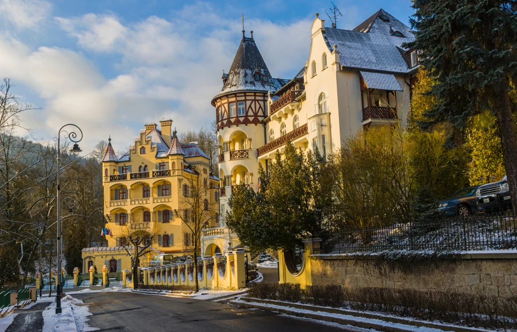 Zabytkowy pałac o żółtej elewacji z fantazyjnymi wieżyczkami i detalami architektonicznymi, osadzony w zimowym krajobrazie z lekko przysypanym śniegiem otoczeniem, przy spokojnej asfaltowej ulicy.