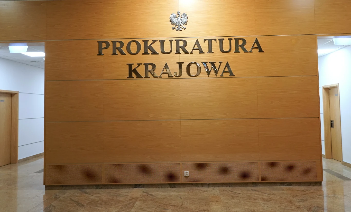 Napis 'Prokuratura Krajowa' na tle drewnianej ściany, powyżej znajduje się godło Polski. W pomieszczeniu widoczna jest błyszcząca podłoga oraz fragmenty drzwi i jasnych ścian po bokach.