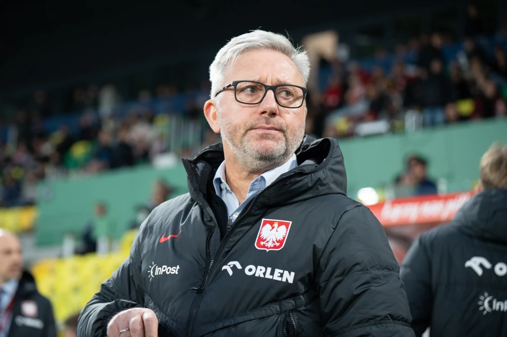 Jerzy Brzęczek Mężczyzna w średnim wieku z siwymi włosami i brodą w okularach, ubrany w czarną kurtkę z logotypami InPost, Orlen oraz godłem Polski. W tle rozmazane trybuny stadionu i widzowie.