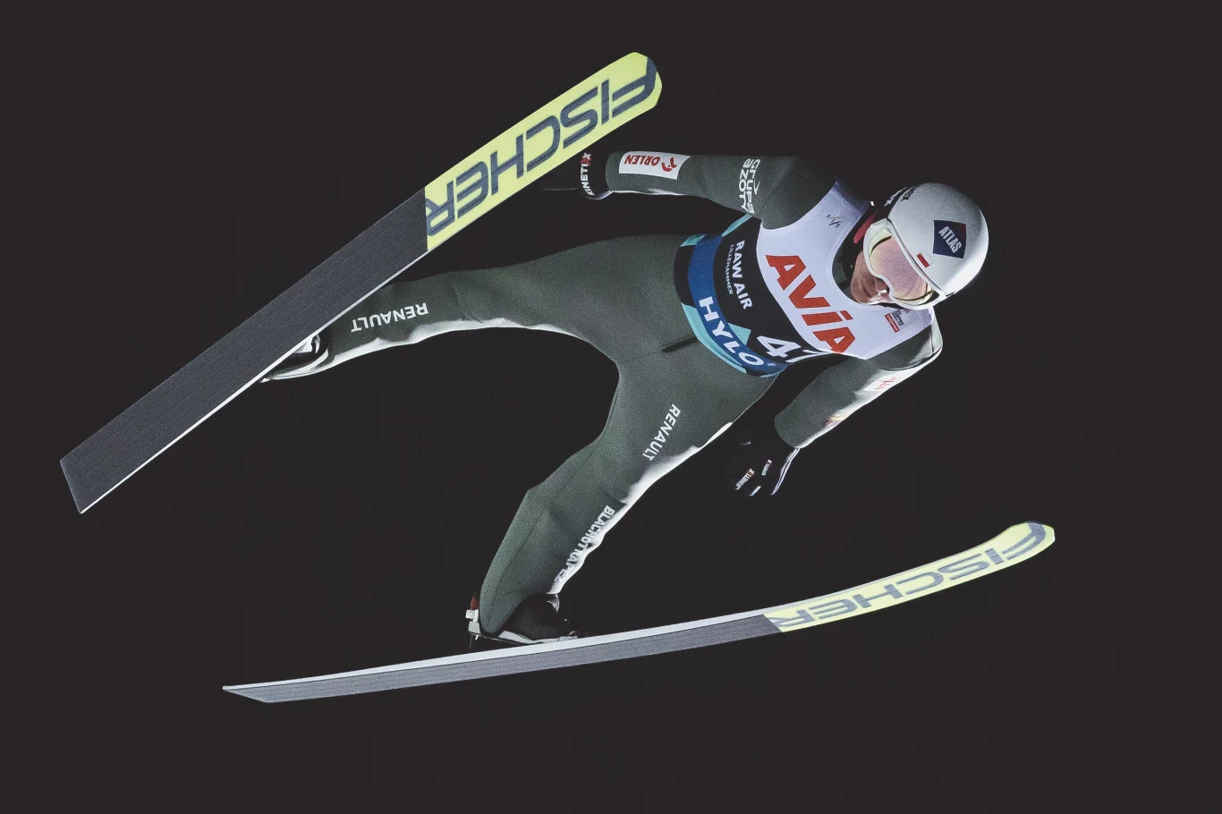 Kamil Stoch w trakcie Pucharu Świata w skokach narciarskich w Lillehammer Zawodnik w powietrzu podczas skoku narciarskiego, pochylony lekko do przodu, w profesjonalnym stroju sportowym i kasku, narty szeroko rozstawione, w tle ciemne niebo.