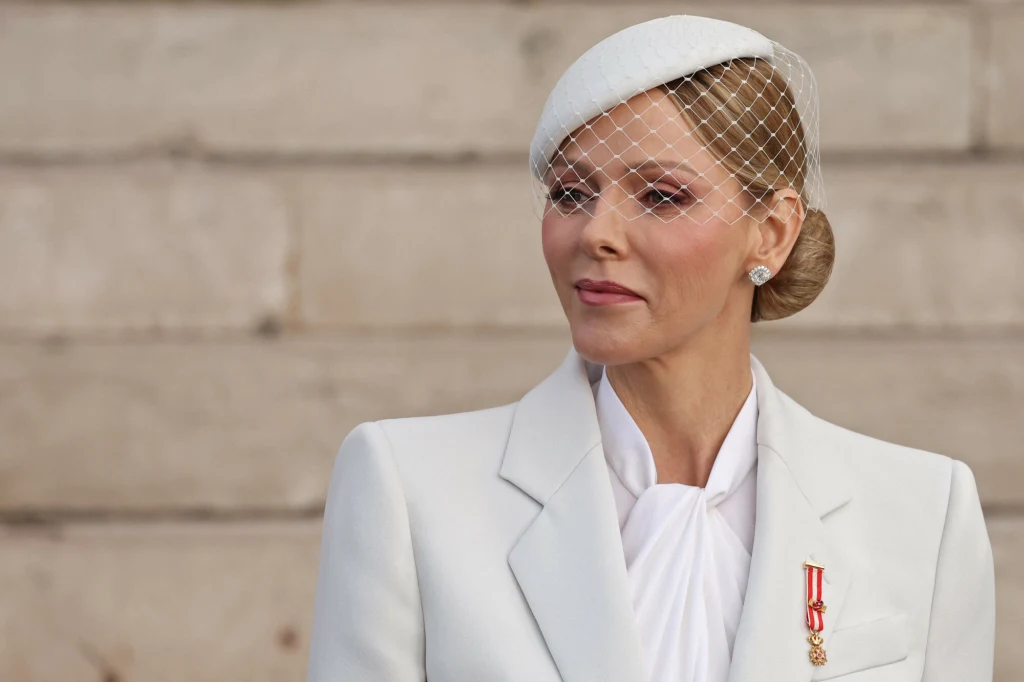 Elegancko ubrana kobieta w białym garniturze z medalem przypiętym do klapy, z jasnym makijażem i upiętymi włosami, na głowie biała toczka z woalką, w tle jasny, kamienny mur.