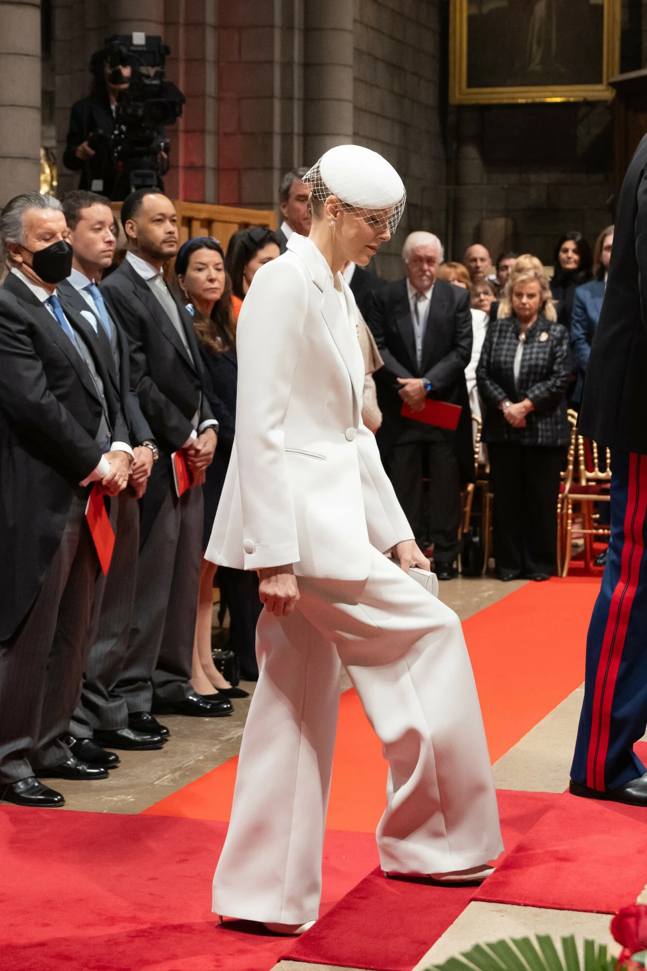 Kobieta ubrana w biały garnitur i biały kapelusz z woalką przechodzi po czerwonym dywanie wśród zgromadzonych oficjalnie ubranych osób podczas uroczystości w reprezentacyjnym, eleganckim wnętrzu.