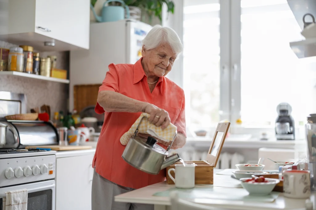 Starsza kobieta nalewa gorącą wodę z czajnika do kubka w jasnej, domowej kuchni. Na blacie znajdują się produkty śniadaniowe, takie jak dżemy i filiżanki, a przez okno wpada naturalne światło.