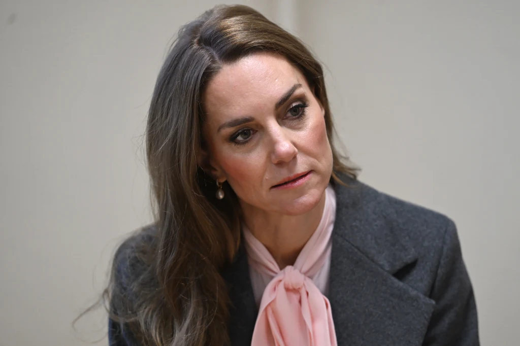 Kobieta o długich, brązowych włosach, ubrana w szary żakiet oraz jasnoróżową bluzkę z kokardą pod szyją, patrząca lekko w bok o poważnym wyrazie twarzy, na jasnym tle.