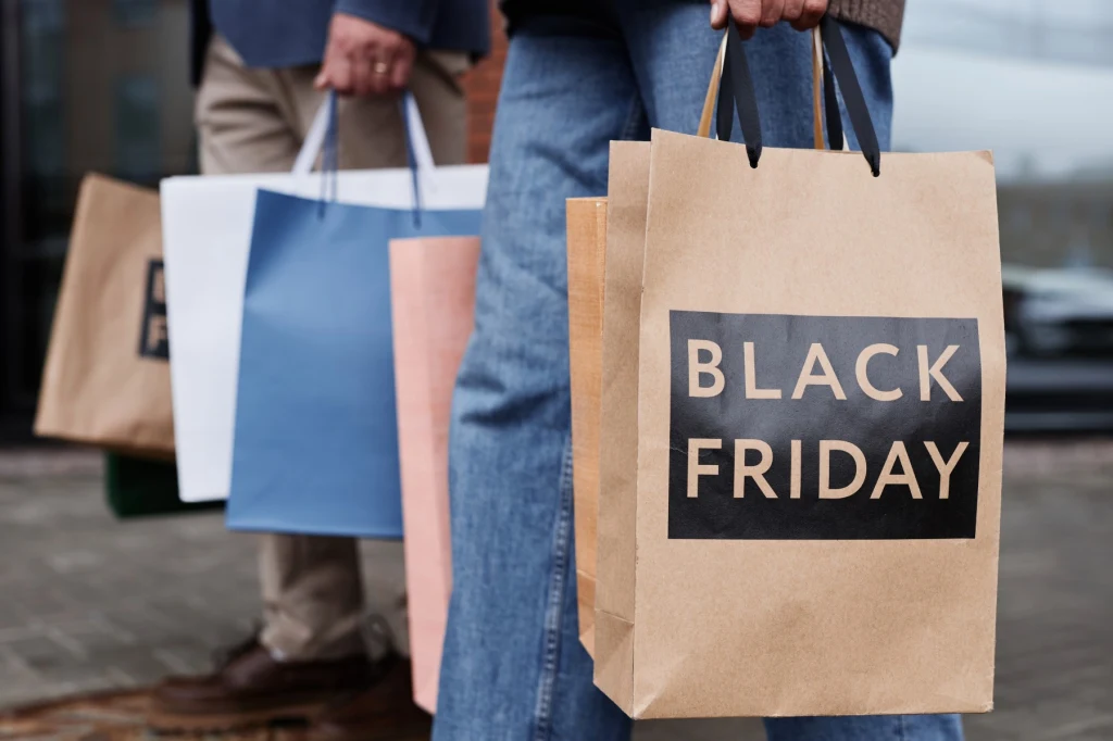 Papierowa torba zakupowa z napisem Black Friday trzymana przez osobę w niebieskich jeansach, w tle widoczne inne torby zakupowe w różnych kolorach trzymane przez drugą osobę.