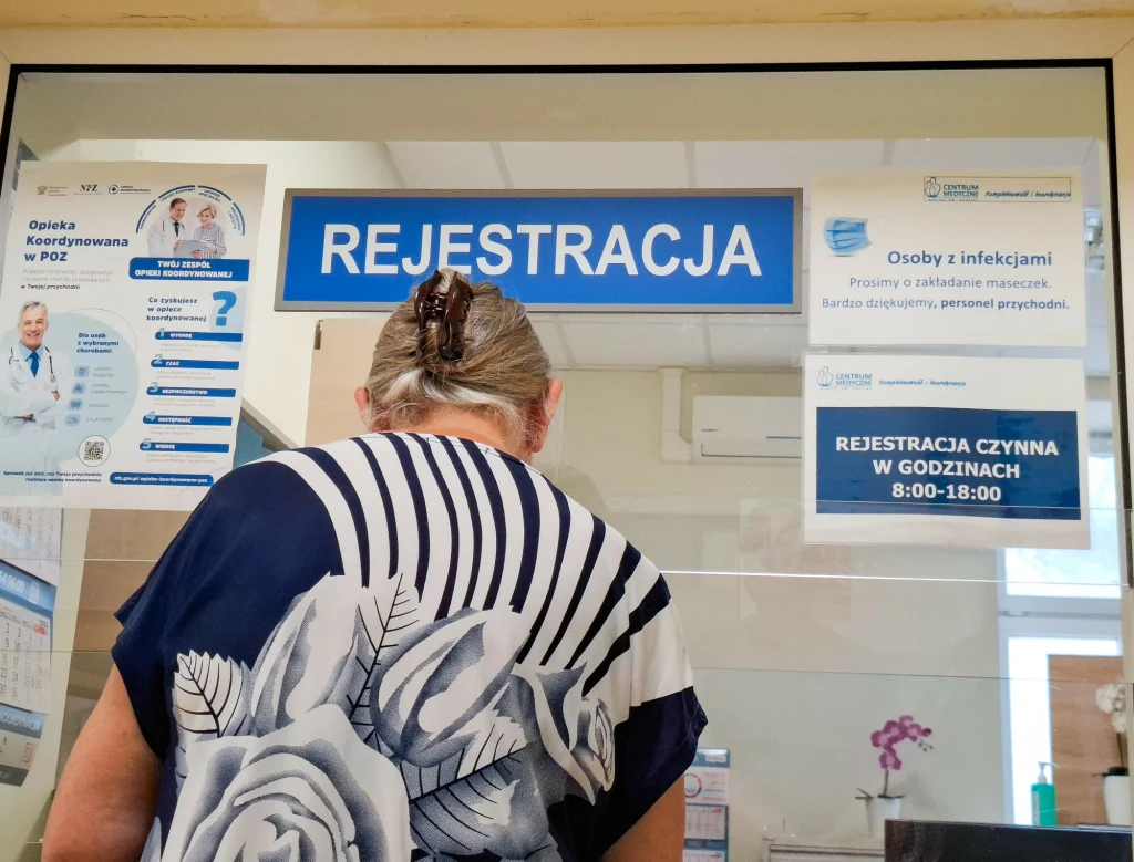 Starsza kobieta w kwiecistej bluzce stoi przed okienkiem rejestracji w przychodni zdrowia. Na ścianie widoczne są informacyjne plakaty dotyczące godzin otwarcia, zaleceń dla osób z infekcjami oraz opieki koordynowanej. Szyba oddziela pacjentów od perso...