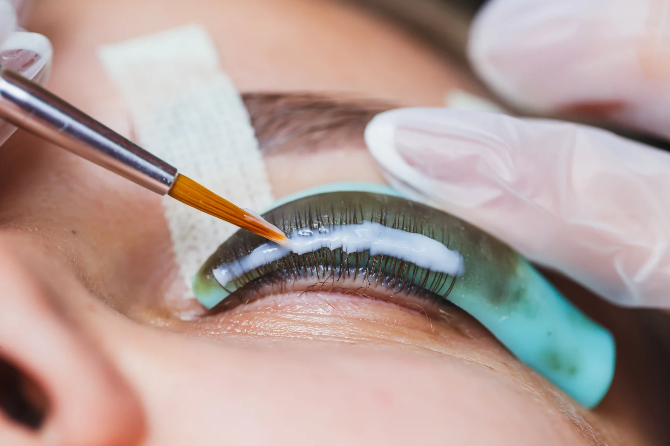 Zbliżenie na zabieg laminacji rzęs, podczas którego specjalistka nakłada pędzelkiem preparat na rzęsy ułożone na silikonowym wałku. Widoczne rękawiczki ochronne oraz opaska zabezpieczająca skórę klientki.
