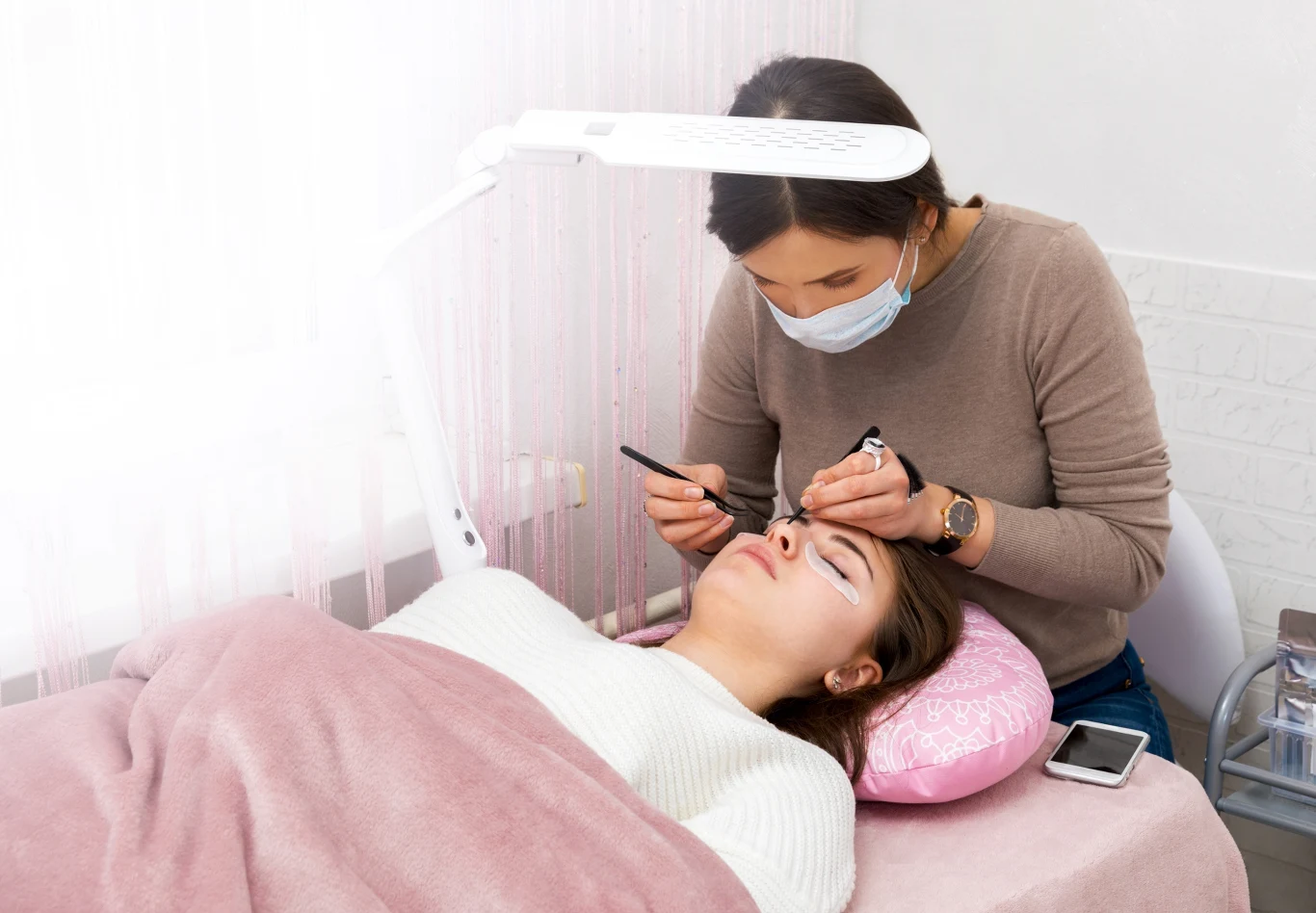 Kosmetyczka wykonuje zabieg przedłużania rzęs klientce leżącej na łóżku zabiegowym, przy użyciu profesjonalnych narzędzi i lampy zabiegowej. Klientka ma zamknięte oczy i zabezpieczoną twarz płatkami ochronnymi.