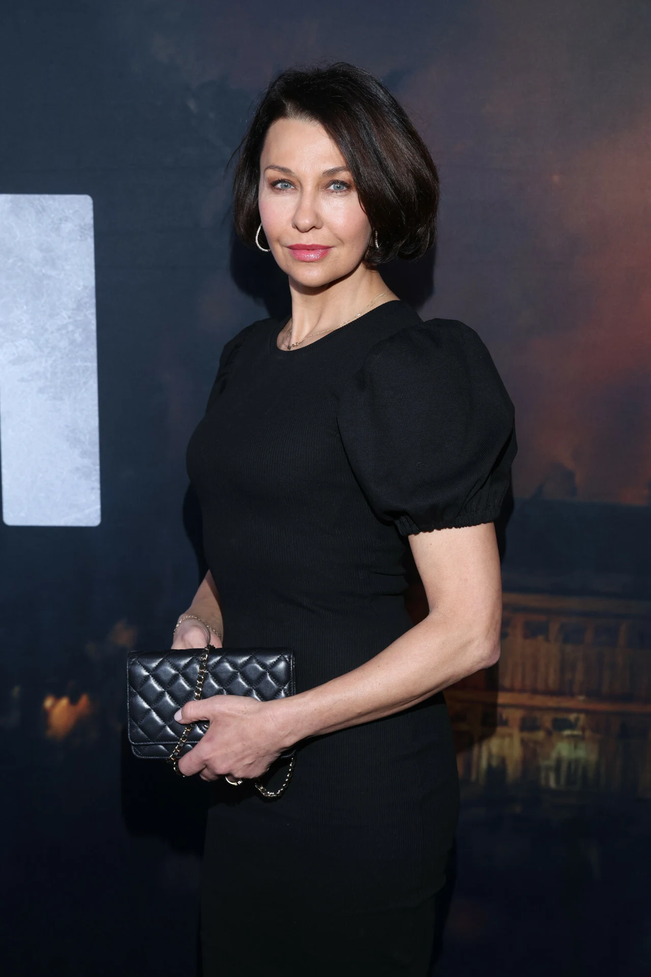 Elegancka kobieta w czarnej sukience, trzymająca w dłoni czarną pikowaną kopertówkę. Spokojny wyraz twarzy, ciemne tło z rozmytymi elementami architektonicznymi i światłami.