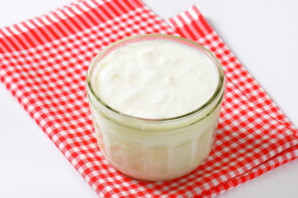 Kefir jest źródłem probiotyków, witamin i minerałów. Warto spożywać go regularnie Szklany słoiczek wypełniony białym, kremowym jogurtem naturalnym ustawiony na czerwono-białej kraciastej ściereczce na jasnym tle.