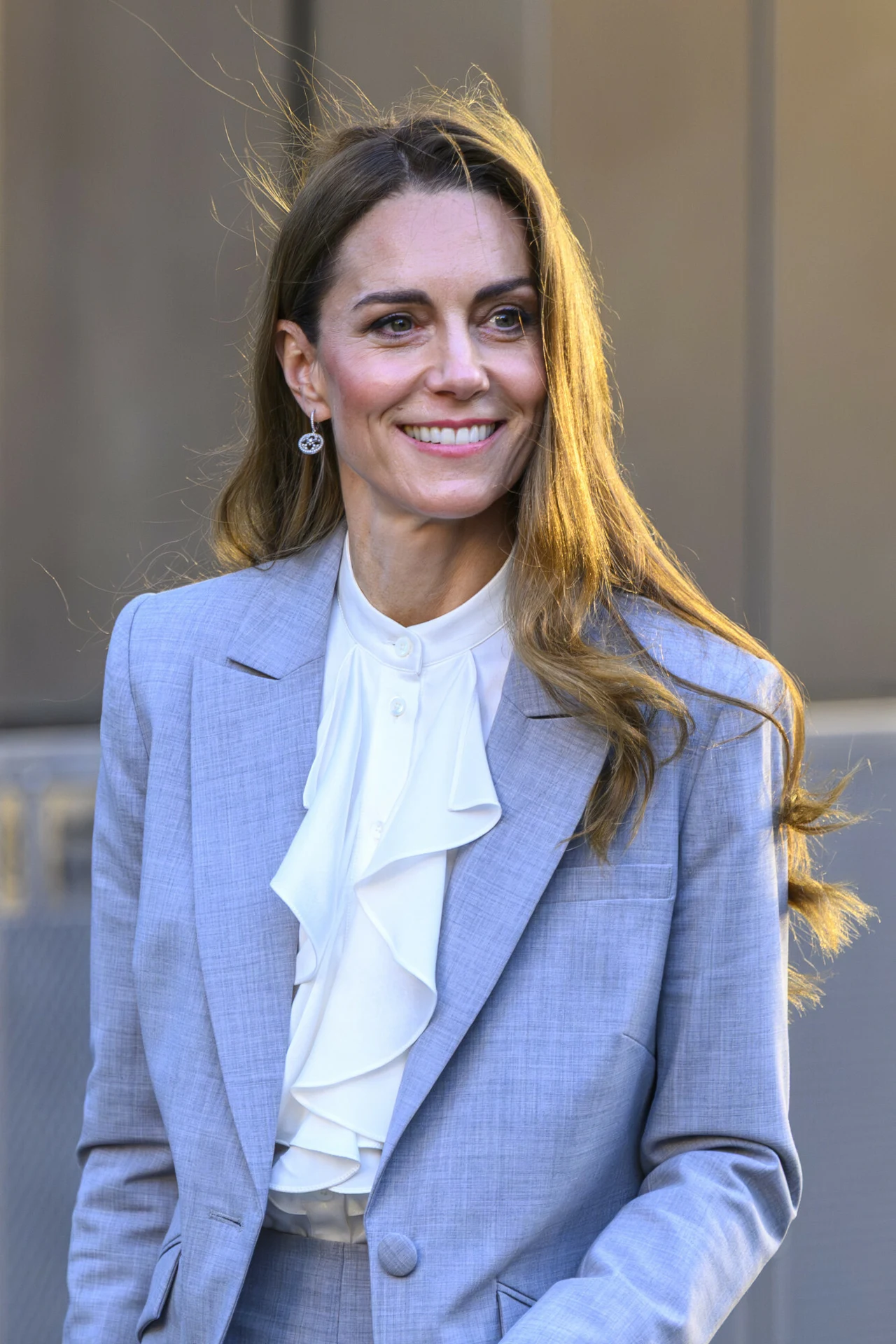 Kobieta z długimi, jasnobrązowymi włosami uśmiecha się, ubrana w jasno-niebieski garnitur z białą, falbanistą koszulą i delikatnymi kolczykami, tło rozmyte, sugerujące miejskie otoczenie.
