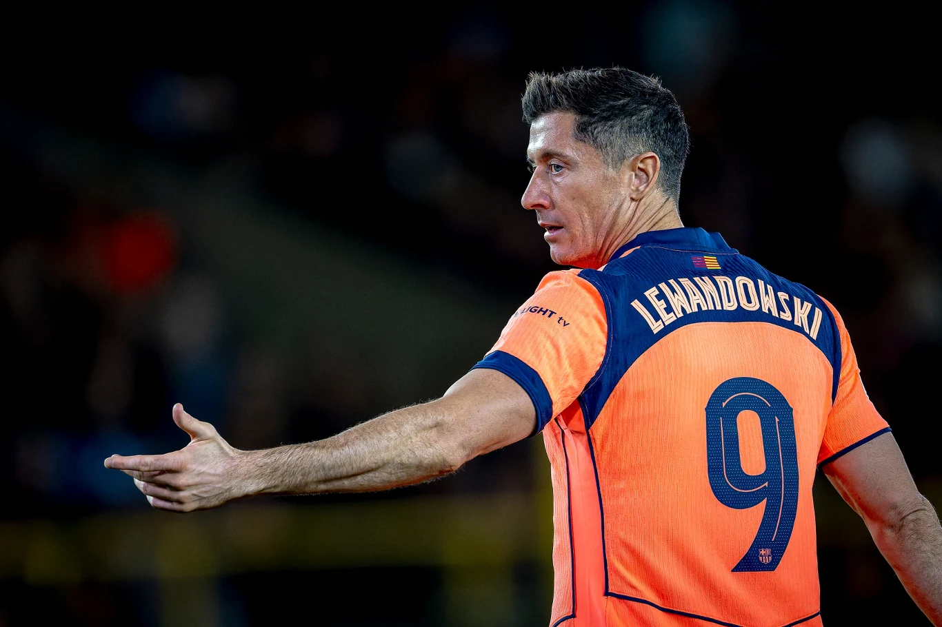 Robert Lewandowski Piłkarz w pomarańczowej koszulce z numerem 9 i nazwiskiem Lewandowski na plecach, wykonujący gest ręką, znajduje się na tle rozmazanego stadionu.