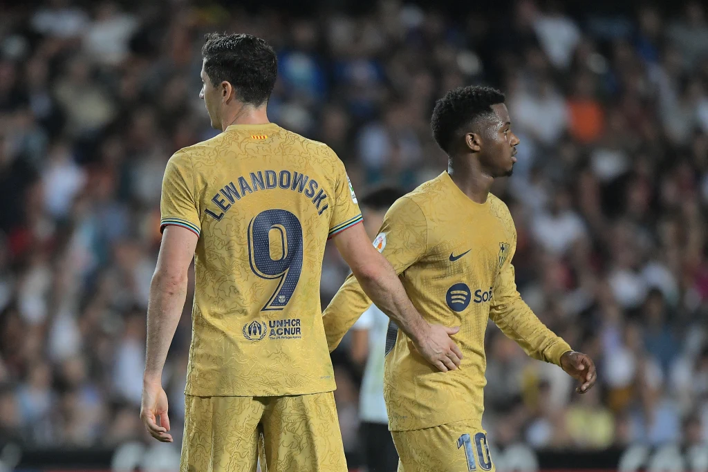 Robert Lewandowski i Ansu Fati Dwóch piłkarzy w złotych strojach drużyny FC Barcelona stoi na boisku, jeden z nich nosi koszulkę z nazwiskiem Lewandowski i numerem 9, drugi z numerem 10. W tle rozmyta publiczność stadionowa.