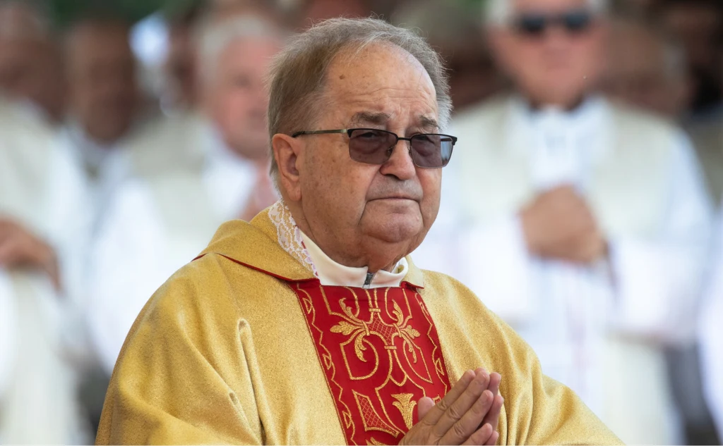 Starszy mężczyzna w okularach ubrany w złotą szatę liturgiczną z czerwonym zdobieniem, dłonie złożone jak do modlitwy, w tle inne osoby ubrane w jasne ornaty.