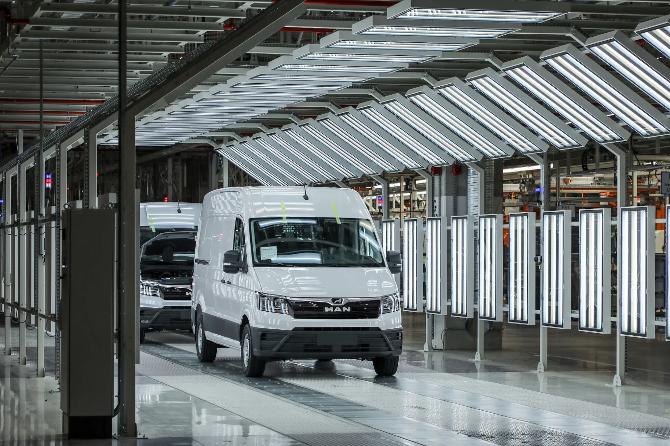 MAN chce zwiększyć produkcję w Polsce Białe samochody dostawcze MAN wyjeżdżają z linii produkcyjnej w hali fabrycznej oświetlonej silnym sztucznym światłem, widoczne liczne lampy nad pojazdami, industrialne otoczenie.