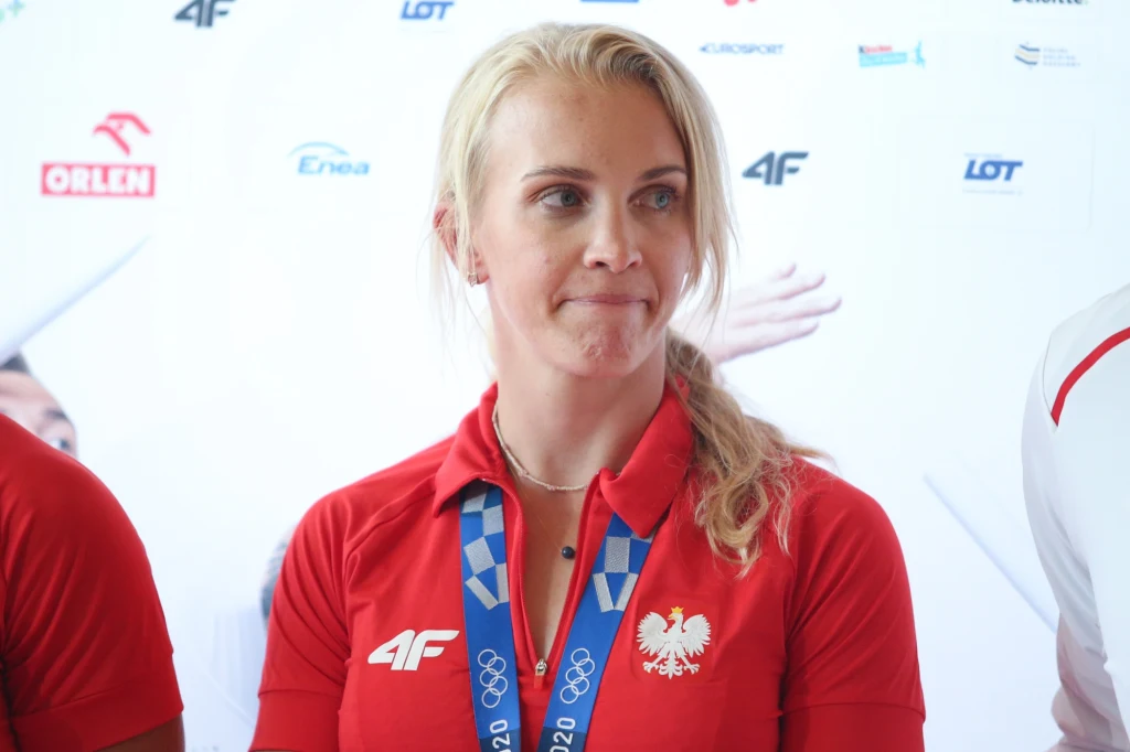 Sportowiec w czerwonej koszulce z godłem Polski i logotypami sponsorskimi, na szyi zawieszony medal olimpijski. Tło z logotypami sponsorów i partnerów, postać patrzy lekko w bok.