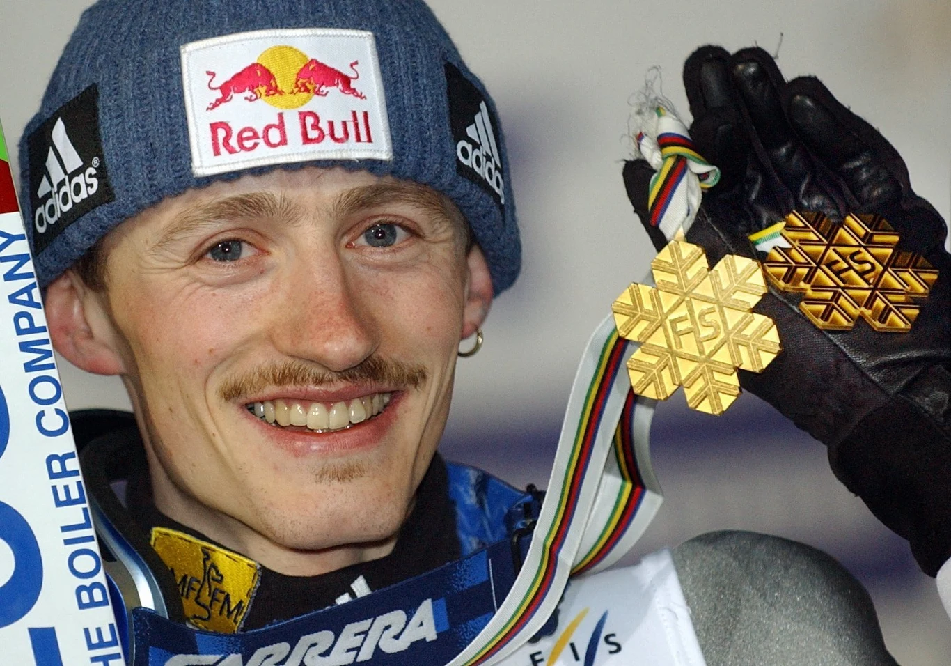 Adam Małysz Uśmiechnięty sportowiec w niebieskiej czapce z logo Red Bull i zimowej kurtce trzyma w dłoni dwa złote medale oraz szarfę z kolorowymi paskami, symbolizującą mistrzostwa świata w narciarstwie.