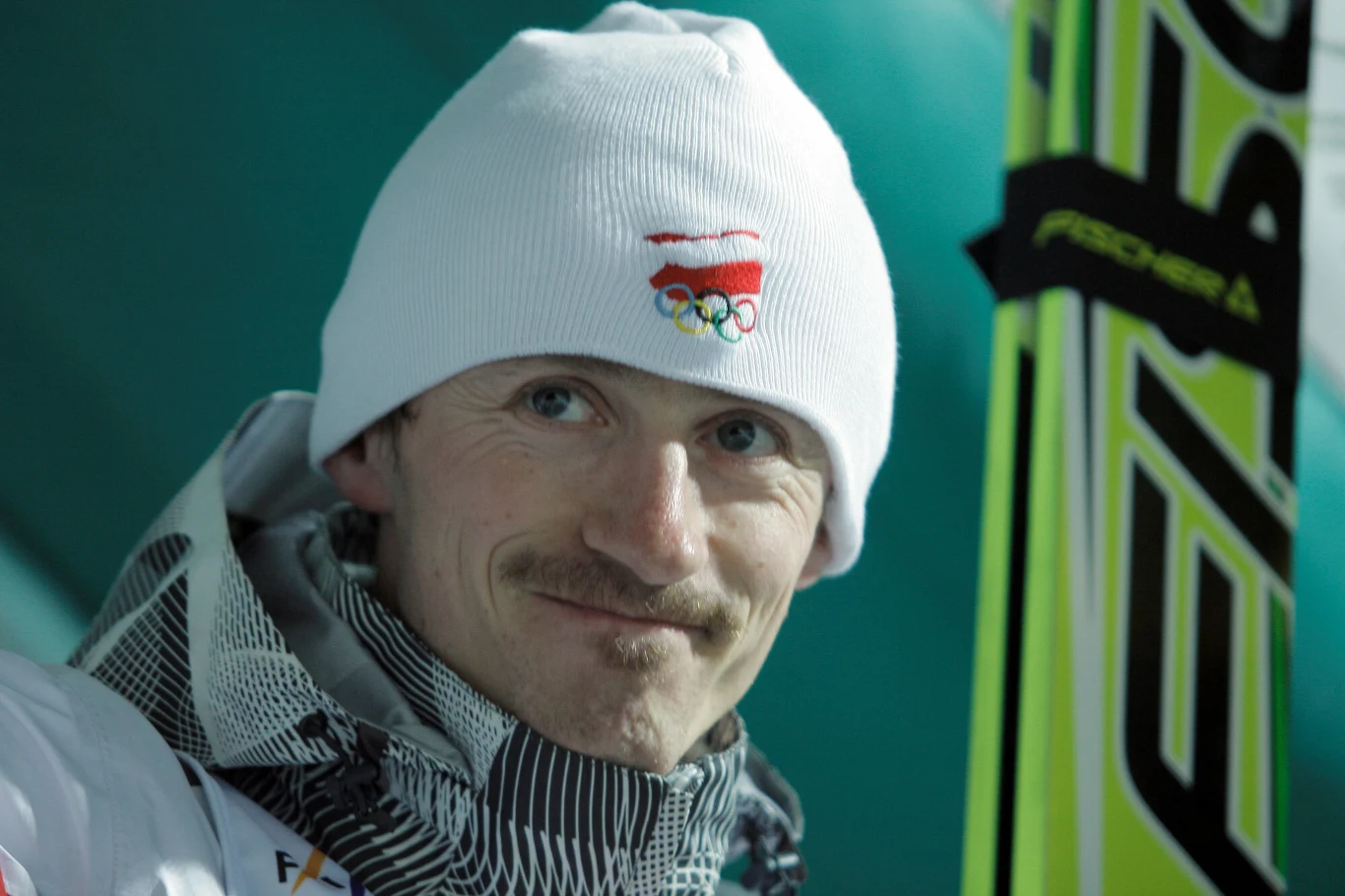 Sportowiec w zimowej czapce z logo olimpijskim, uśmiechnięty, trzymający narty Fischer na jasnozielonym tle inspirowanym sportami zimowymi.