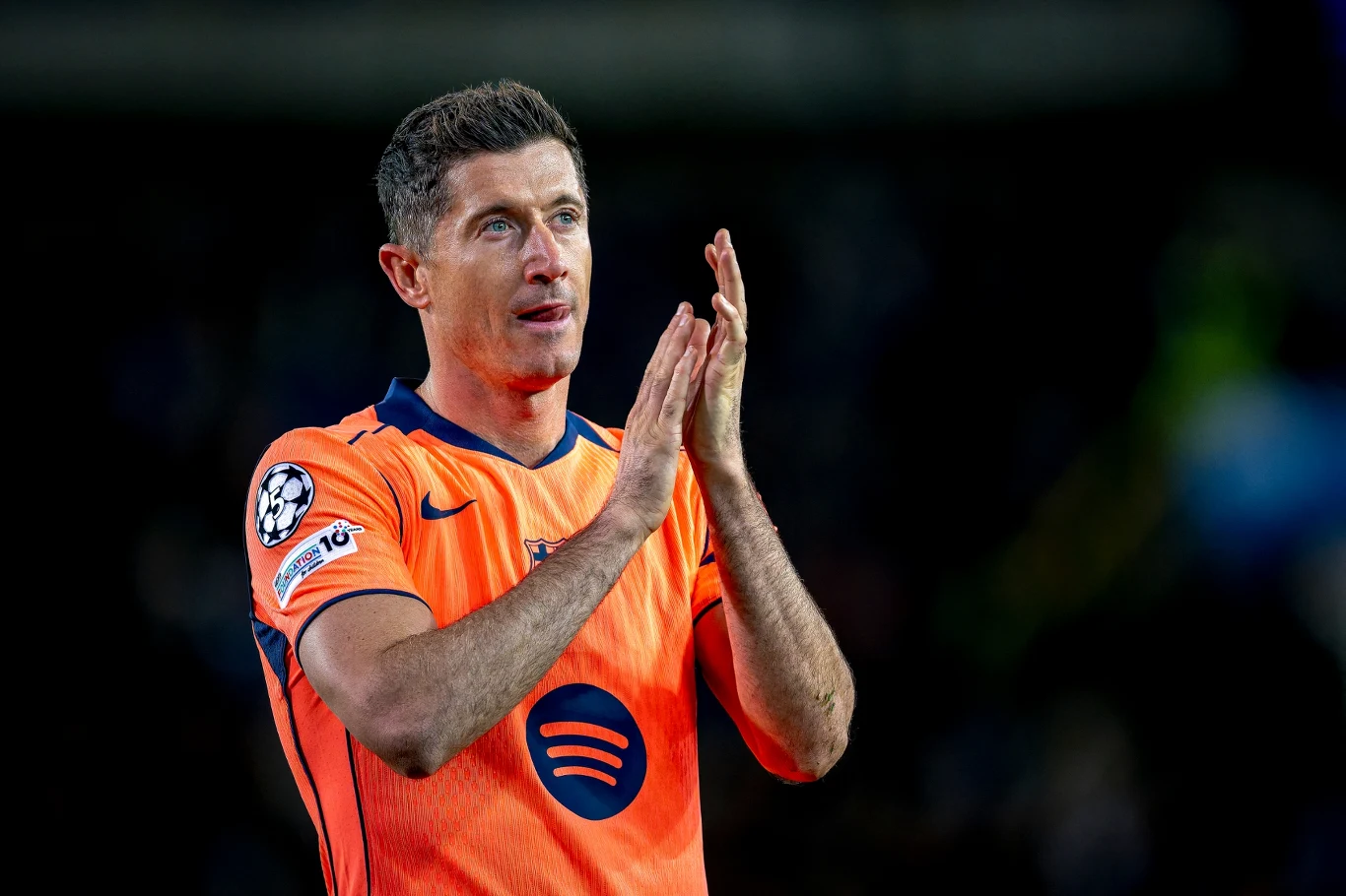 Robert Lewandowski Piłkarz w pomarańczowym stroju FC Barcelony z widocznym logotypem Spotify, wykonujący gest brawa na tle rozmytego stadionu