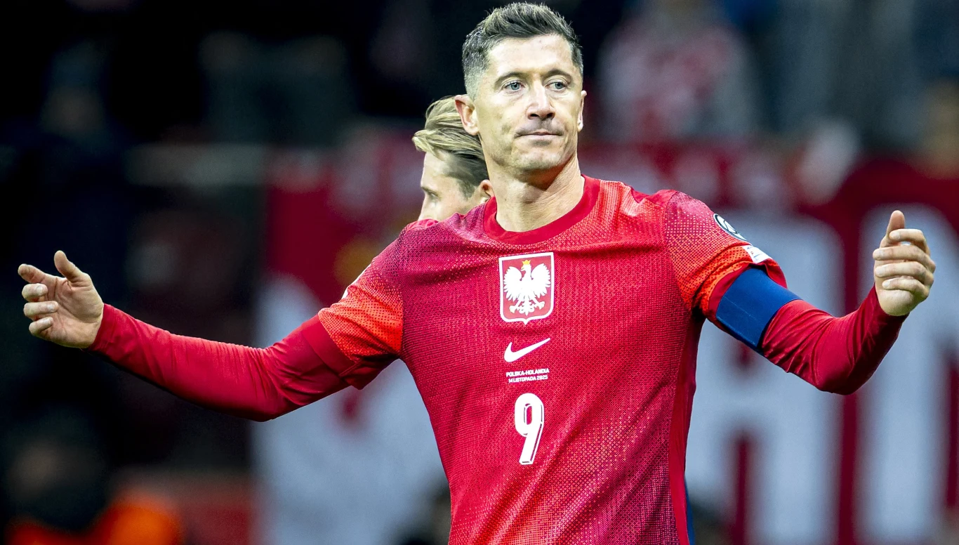 Piłkarz w czerwonej koszulce z orłem i numerem 9 na piersi podczas meczu, unosi ręce w geście otwartości. W tle widoczna rozmyta publiczność stadionu.