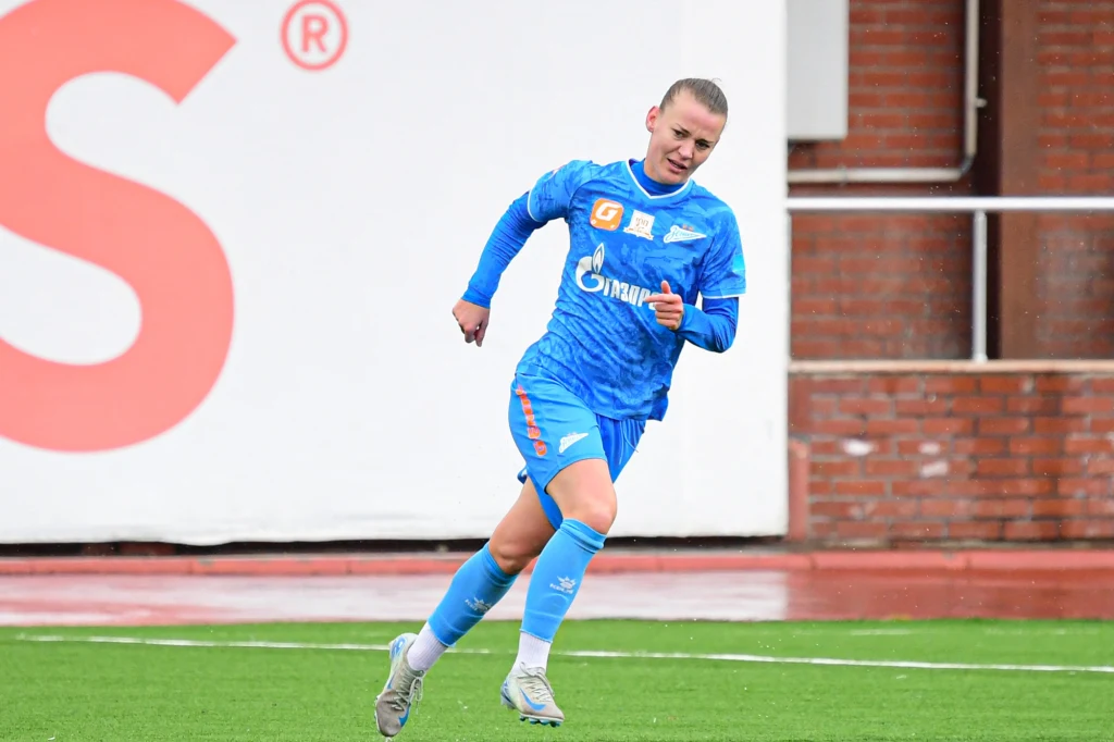 Piłkarka w niebieskim stroju sportowym biegnie po zielonej murawie, w tle widoczny jest fragment boiska oraz ceglana ściana.